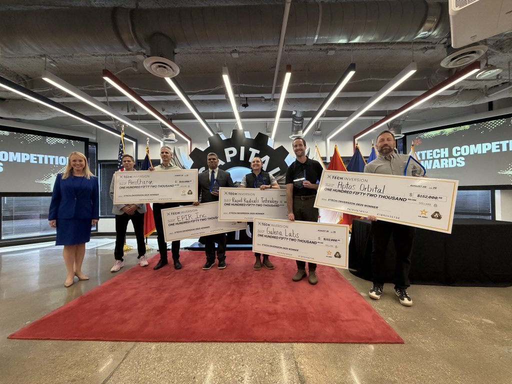 Individuals standing in convention center on red rug with large checks in hand.